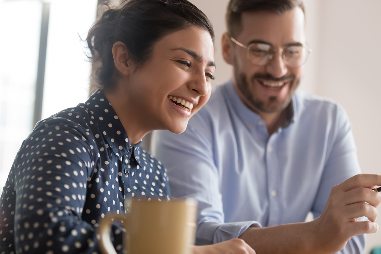 Happy young smiling diverse couple of female indian and male caucasian coworkers watching funny videos during coffee break, getting positive news, making good deal, holding video call with client